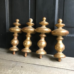 May include: Four antique gold-painted wooden finials with a decorative design. The finials are arranged in a row on a wooden surface.