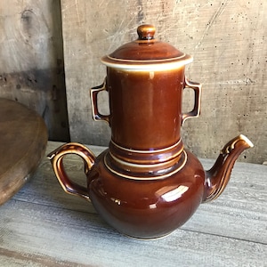 May include: A brown ceramic coffee pot with a lid and a spout. The pot has a white band around the top and bottom.