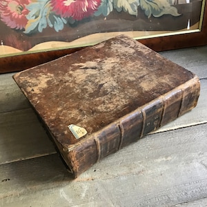 Op de afbeelding: Een vintage, bruin lederen gebonden boek met een versleten en verweerde uitstraling. Het boek heeft een rug met verhoogde banden en is gesloten. Het boek ligt op een houten oppervlak.