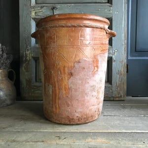 May include: A large, antique terracotta pot with a wide cylindrical shape. The pot has a textured surface with carved geometric patterns and two small handles. The color is a warm, earthy brown with areas of lighter, faded tones, suggesting age and use.