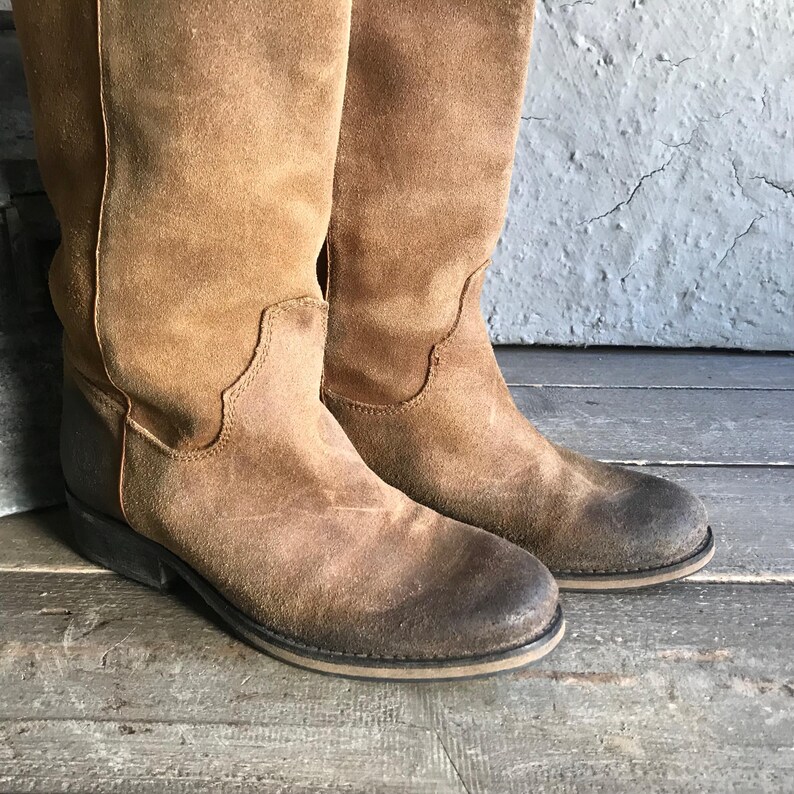 May include: A pair of brown suede knee-high boots. The boots have a worn, distressed appearance with a slightly faded color. The soles are dark brown with a light-colored trim. The boots are displayed on a wooden surface.
