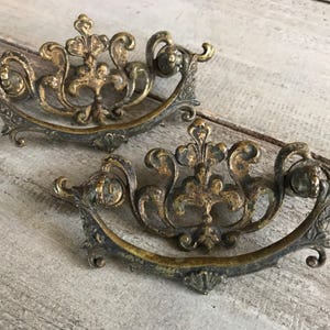 May include: Two ornate, antique-style, brass drawer pulls with intricate floral designs. The pulls are in the shape of a half-circle with a decorative, raised center.