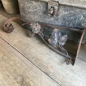 May include: A rustic metal shelf bracket with a decorative grapevine design. The bracket is made of dark brown metal and features a curled end and a grape cluster.