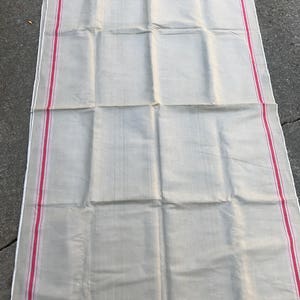 以下が含まれることがあります： 縁に赤いストライプが入った、大きなオフホワイトのリネン製のテーブルクロス。テーブルクロスは半分に折り畳まれており、わずかに質感のある表面をしています。