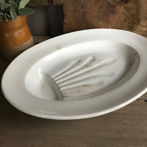 Puede incluir: Plato de cerámica blanca con un diseño elevado y acanalado en el centro. El plato tiene forma ovalada y un borde ligeramente elevado.