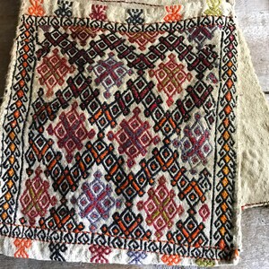 May include: A white fabric square with a colorful geometric pattern embroidered in red, orange, yellow, green, blue, black, and purple. The fabric has a rough texture and is fringed on all sides.