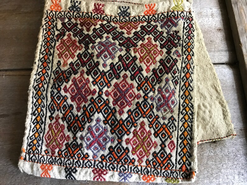May include: A white fabric square with a colorful geometric pattern embroidered in red, orange, yellow, green, blue, black, and purple. The fabric has a rough texture and is fringed on all sides.