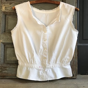 May include: A sleeveless white blouse featuring a button-down front, a round neckline with delicate lace trim, and a cinched waist with vertical pleats. The garment includes small red embroidered letters H C and the number 42 near the waistline.