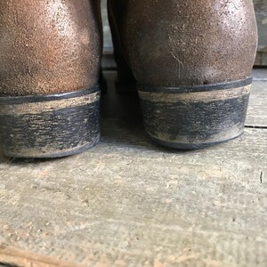 May include: Close-up of the heels of two brown leather boots. The heels are black with a worn, distressed appearance. The boots appear to be well-worn, with visible signs of use and age. The boots are on a wooden surface.