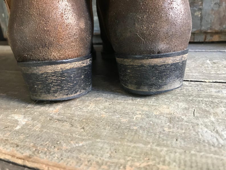 May include: Close-up of the heels of two brown leather boots. The heels are black with a worn, distressed appearance. The boots appear to be well-worn, with visible signs of use and age. The boots are on a wooden surface.