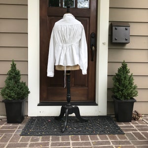 May include: White cotton blouse with long sleeves and a gathered back. The blouse is cinched at the waist with a white drawstring.