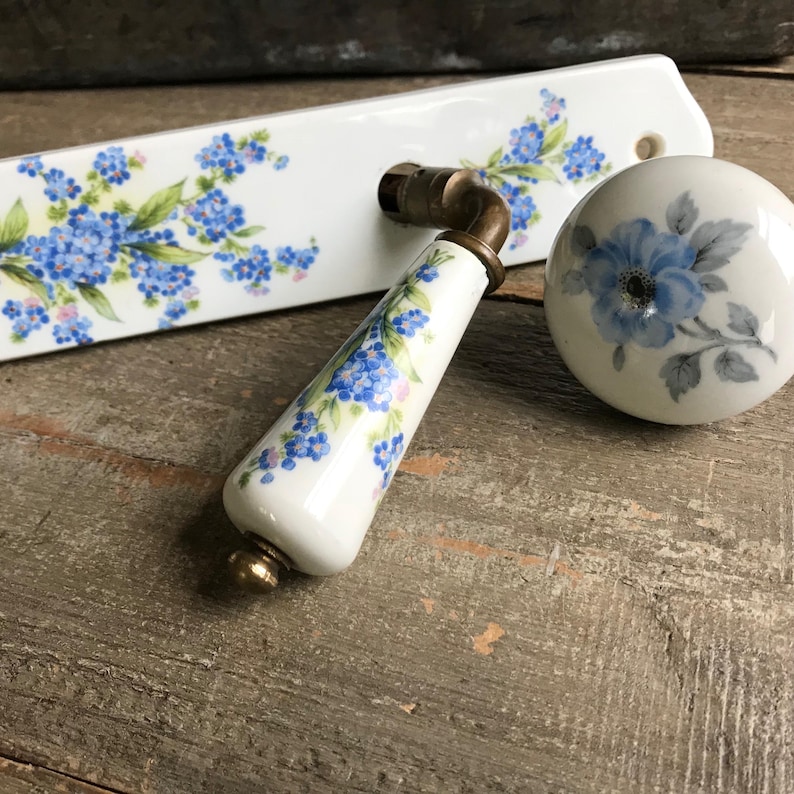 May include: A white porcelain door handle set with a floral design. The handle is decorated with blue forget-me-not flowers and green leaves. The knob is also decorated with a blue forget-me-not flower.
