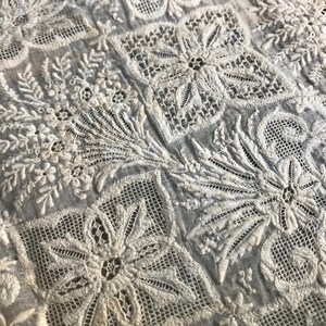 以下が含まれることがあります： 繊細なレースの質感を持つ、白い綿生地に複雑な刺繍の花柄が施されています。花と葉の繰り返しパターンが特徴です。
