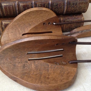 May include: Pair of antique wooden shoe stretchers with metal hardware. The stretchers are a warm brown color and have a curved shape with slots. The metal components are a dark, aged metal. The background includes antique books.