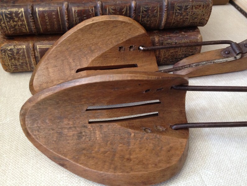 May include: Pair of antique wooden shoe stretchers with metal hardware. The stretchers are a warm brown color and have a curved shape with slots. The metal components are a dark, aged metal. The background includes antique books.