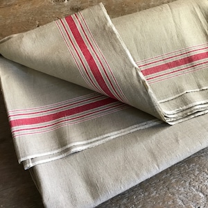 Pode incluir: Um tecido de linho dobrado com listras vermelhas e brancas. O tecido é de cor bege natural e tem um visual rústico e vintage.