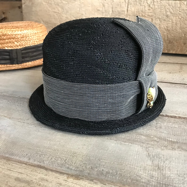 May include: A black and white straw hat with a wide brim and a black ribbon. The hat has a small gold and green embellishment on the side.