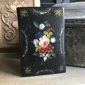 May include: A black wooden box with a floral design painted on the front. The flowers are painted in shades of pink, red, blue, and white, and are accented with iridescent mother-of-pearl accents. The box is a vintage piece and has a distressed look.