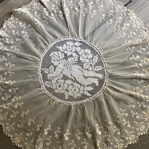 Puede incluir: Un mantel de encaje blanco con un diseño circular que presenta dos querubines sosteniendo flores. El mantel tiene un borde festoneado y está decorado con pequeños motivos florales.