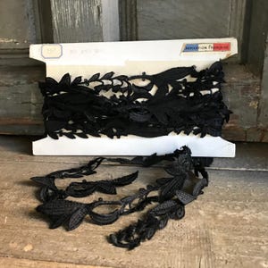 Puede incluir: Ribete de encaje floral negro sobre una bobina de cartón blanco. El ribete presenta un diseño de hojas detallado con pequeños detalles de cuentas. La bobina tiene el texto "ABRICATION FRANCAISE".