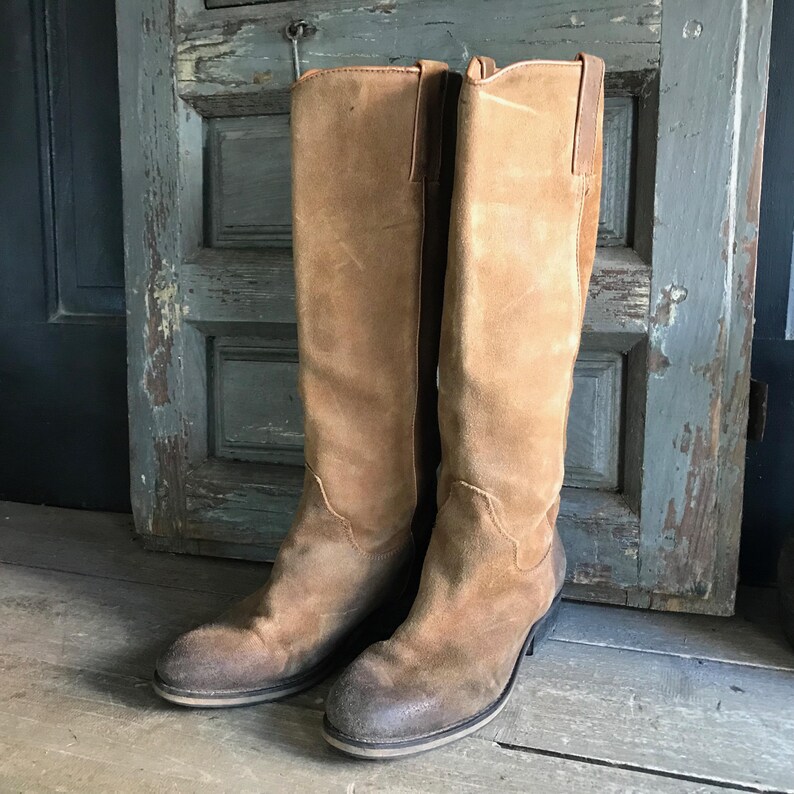 May include: A pair of tall, tan suede boots with a worn appearance. The boots have a classic design, featuring a rounded toe, a mid-height heel, and a tall shaft that extends to the knee. The boots are set against a weathered wooden background.