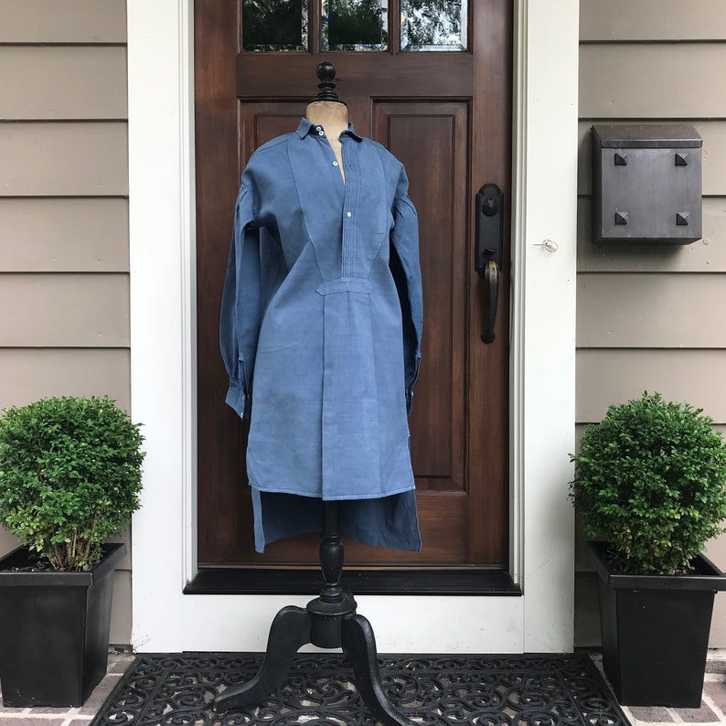 May include: A blue linen shirt with a long, pointed collar and a button-up front. The shirt has a long, flowing hemline and is displayed on a black mannequin.