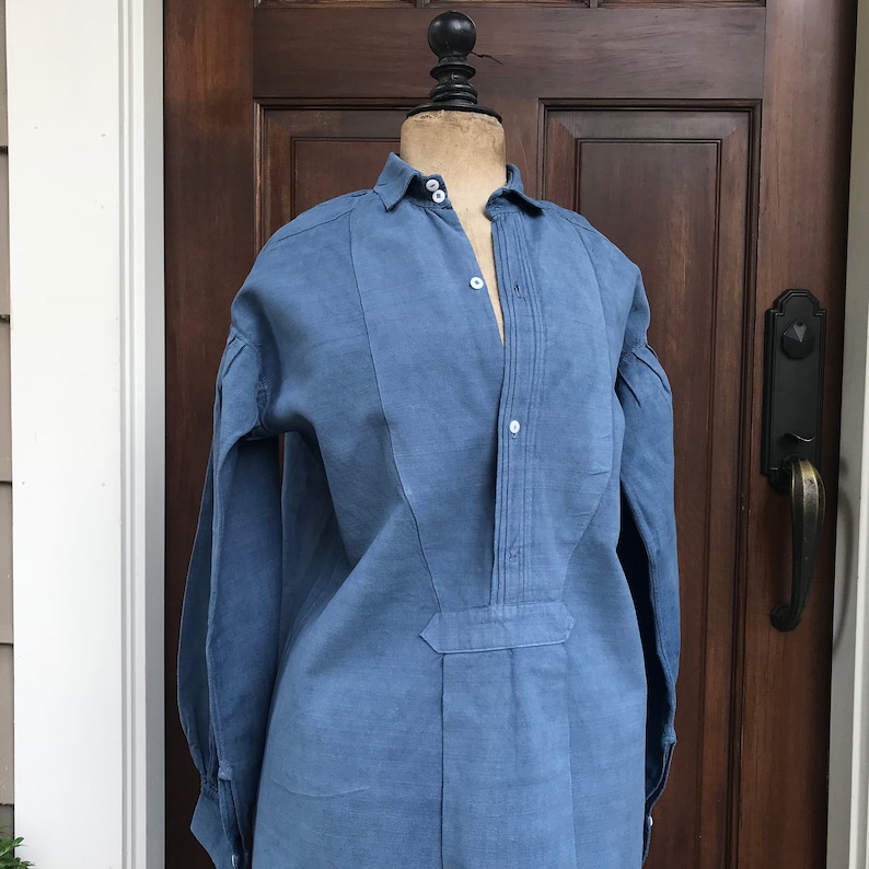 May include: A blue linen shirt with a white collar and button-up front. The shirt has a pleated detail down the front and long sleeves.