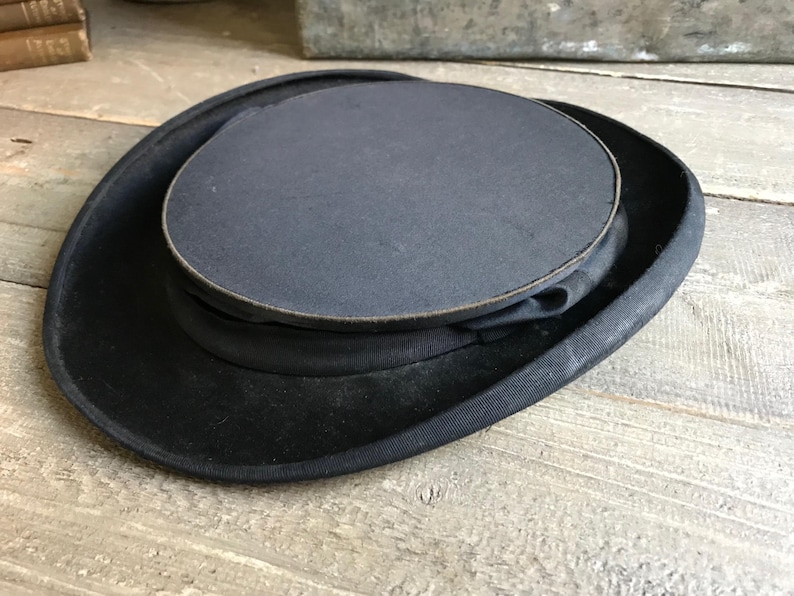 May include: A black top hat with a wide brim and a tall, cylindrical crown. The hat is made of felt and has a smooth, polished finish.