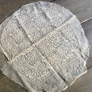 以下が含まれることがあります： 繊細な花柄の白い丸いレースのテーブルクロス。テーブルクロスは縁がほつれていて、少しすり切れています