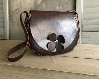 Puede incluir: Un bolso bandolera de cuero marrón con un detalle floral. La flor está hecha de cuero y tiene una gema turquesa en el centro.
