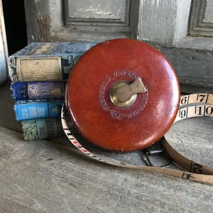 Puede incluir: Una cinta métrica vintage, redonda, de color marrón rojizo, con un mecanismo de enrollado de latón. La cinta está parcialmente extendida, mostrando marcas de medición. Una pila de libros antiguos pequeños es visible en el fondo, sugiriendo un tema de historia o coleccionables.