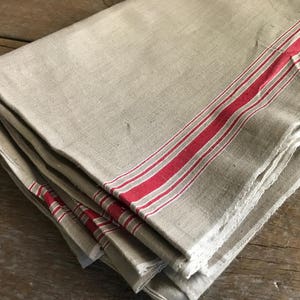 以下が含まれることがあります： 赤いストライプの入ったリネン製のキッチンタオルの束。タオルは折り畳まれており、素朴でヴィンテージな外観をしています。