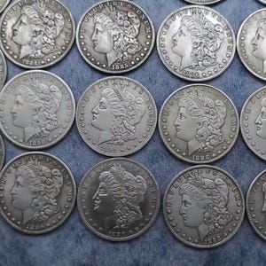 May include: A collection of silver-toned Morgan dollar coins arranged in a grid. Each coin features the profile of Lady Liberty facing left, surrounded by the text E PLURIBUS UNUM and the year of minting, including dates from 1879 to 1898.