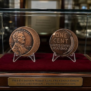 Può includere: Due monete color rame esposte in una teca di vetro su una base di velluto rosso. La moneta a sinistra mostra Abraham Lincoln e l'anno 1914. La moneta a destra riporta il testo ONE CENT UNITED STATES OF AMERICA. Una targhetta recita 1914-D LINCOLN WHEAT CENT OFF-CENTER ERROR COLLECTOR'S EDITION PREMIUM REPRODUCTION.