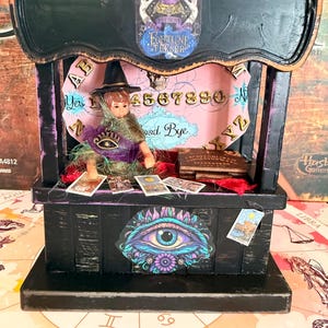 May include: A black wooden fortune teller booth featuring a miniature figure in a witch hat, a spirit board backdrop with numbers and letters, tarot cards, a small book, and a central eye graphic. The top arch displays a crystal ball illustration.