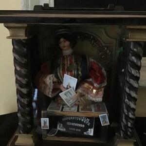 May include: A wooden box with a glass front, containing a doll dressed in a red and blue costume holding tarot cards. The box also contains a Ouija board with the letters "NOPQRS TUVWXYZ 1234567890 GOOD BYE".