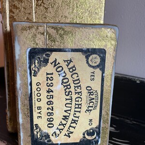 May include: A gold-painted wooden bookend shaped like a book with a vintage Ouija board design. The board features the letters A-Z, numbers 0-9, and the words "YES", "NO", and "GOOD BYE".