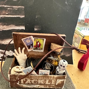 May include: A wooden box labeled "TACKLE" filled with occult items. Includes tarot cards, a skull, a hand, a bottle with a cork, and a small skull figurine. The box is brown with a brown cord.
