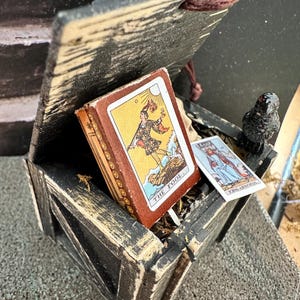 May include: A small, weathered wooden box with a hinged lid, containing tarot cards. The cards feature colorful illustrations and the words "The Fool" and "King of Swords." A small, black bird figurine sits on the side of the box.