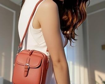 Puede incluir: Un bolso bandolera de color terracota con acabado texturizado, que presenta un bolsillo frontal con solapa asegurado por una correa vertical y una hebilla de metal plateado. El bolso incluye una correa de hombro ajustable a juego y cremallera.