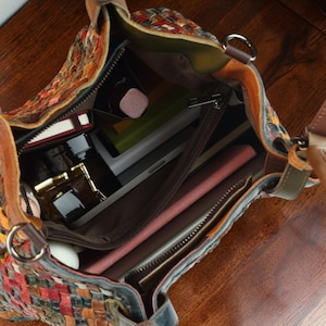 Puede incluir: Un bolso de mano de cuero trenzado multicolor con apertura superior, que cuenta con un forro interior marr&oacute;n, un compartimento central con cremallera y varios objetos personales en su interior, incluyendo un objeto rectangular rosa y recipientes.