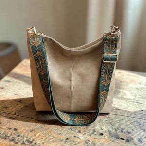 Puede incluir: Un bolso hobo de color beige con una costura central vertical y una correa ajustable para el hombro. La correa está decorada con un patrón geométrico en azul verdoso, naranja y dorado e incluye herrajes metálicos dorados.