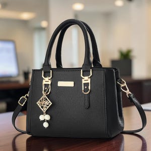 Puede incluir: Un bolso de mano negro con textura, dos asas superiores y una correa de hombro extraíble. El bolso cuenta con herrajes en tono dorado, una placa metálica rectangular en la parte frontal y un colgante decorativo con marco geométrico y tres cuentas blancas.