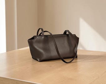 Puede incluir: Un bolso de hombro de color marrón oscuro con un acabado liso y mate. El bolso presenta una base rectangular, una abertura superior ligeramente curvada y dos correas finas a juego fijadas a los bordes superiores del cuerpo principal.