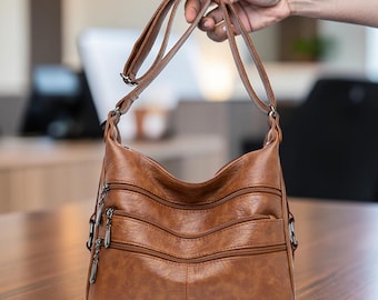 Puede incluir: Un bolso bandolera marrón de textura lisa, con correa ajustable y tres compartimentos horizontales con cremallera en la parte frontal. Cada cremallera tiene un tirador metálico y el bolso cuenta con una base redondeada y estructurada.