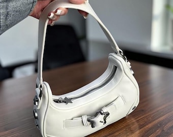Puede incluir: Un bolso de hombro blanco en forma de media luna con cierre de cremallera superior con tirador en forma de estrella, una correa decorativa en la parte delantera con hebilla de estrella y detalles metálicos de estrellas en los lados.