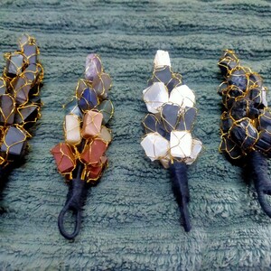 May include: Four decorative wall hangings shaped like feathers, each crafted from clusters of raw gemstones wrapped in gold-colored wire. The stems are wrapped in black thread with a loop at the base. The stones include dark gray, multicolored, white, and black varieties.