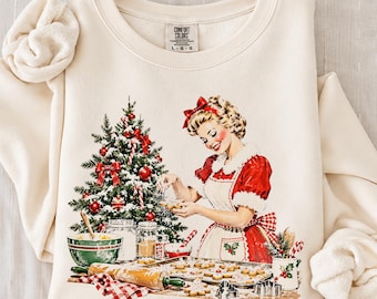 Könnte beinhalten: Ein cremefarbenes Sweatshirt mit Rundhalsausschnitt und einem Grafikdruck einer Person in einem rot-weißen Kleid, die neben einem geschmückten Weihnachtsbaum Plätzchen backt. Das Etikett am Halsausschnitt zeigt Comfort Colors L G G.