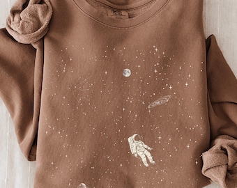 Könnte beinhalten: Ein braunes Langarm-Sweatshirt mit Rundhalsausschnitt und einem weißen Grafikdruck eines Astronauten, der im Weltraum zwischen Sternen, einem Planeten und einem Himmelskörper mit Ringen schwebt. Der Stoff ist weich mit gerippten Bündchen.