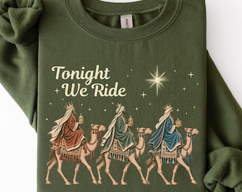 Könnte beinhalten: Ein olivgrünes Sweatshirt mit Rundhalsausschnitt und einer Grafik von drei auf Kamelen reitenden Figuren, darüber der Text Tonight We Ride und ein Stern. Die Figuren tragen rote, grüne und blaue Gewänder und halten jeweils ein goldenes Objekt.